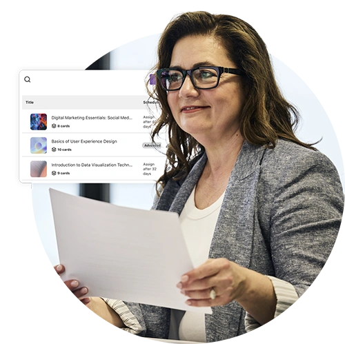 A woman with glasses holding a paper and smiling, with a digital screen showing course titles behind her.