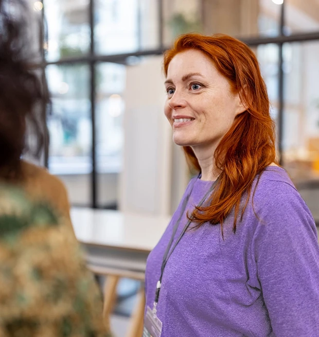 A woman with long red hair wearing a purple shirt and a lanyard talks and smiles in a bright space.