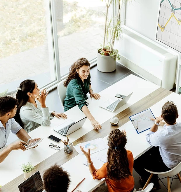 A diverse group of six people in a meeting, discussing over laptops and charts in a bright room.