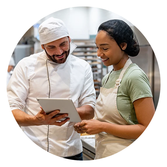 A chef and a female baker smile while looking at a tablet together in a bakery.