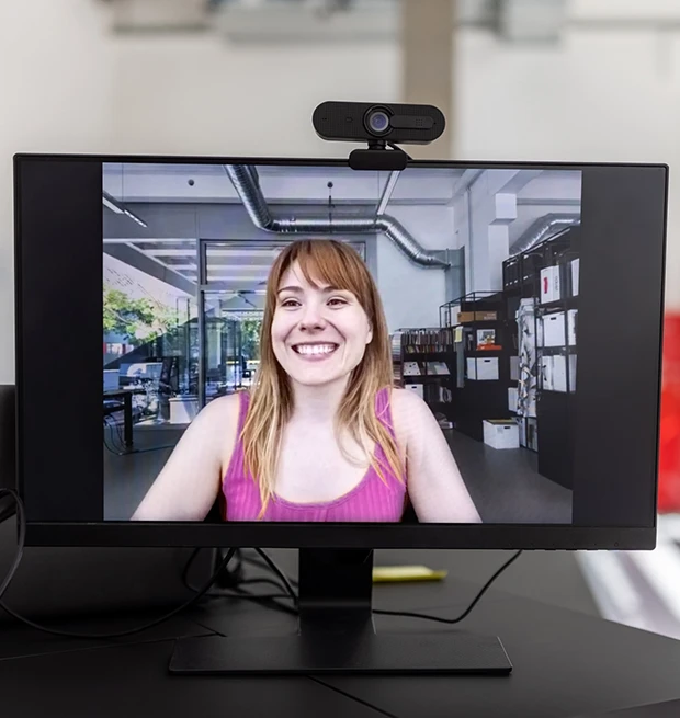 A smiling woman with long hair in a pink tank top is on a computer screen, set in a modern office.