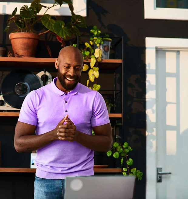 A man in a purple polo shirt smiles while standing in front of a laptop, surrounded by plants.