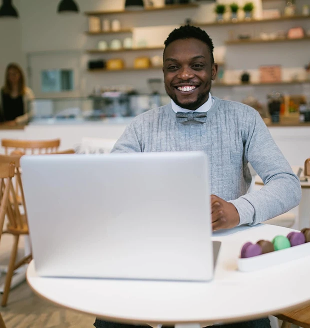 A smiling man in a gray sweater and bow tie sits at a table with a laptop, overlooking colorful candies.