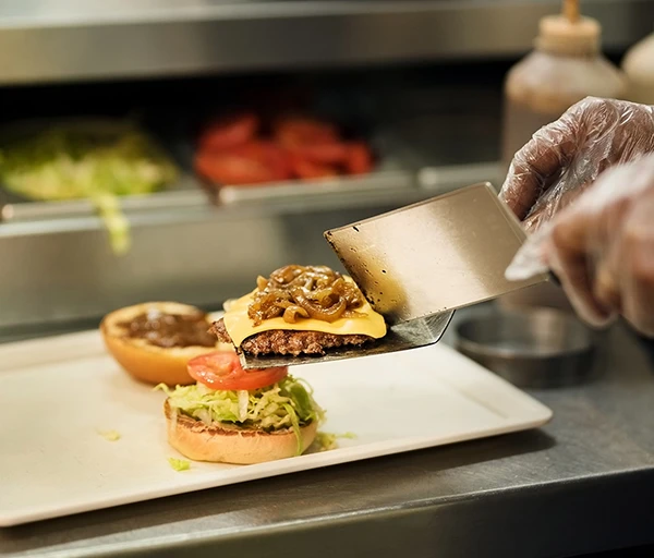 A hand with a spatula lifts a cheeseburger with onions, ready to place on a bun with lettuce and tomato.
