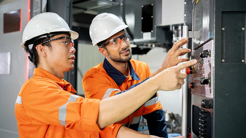 Two factory workers in hard hats showing each other controls on a machine.