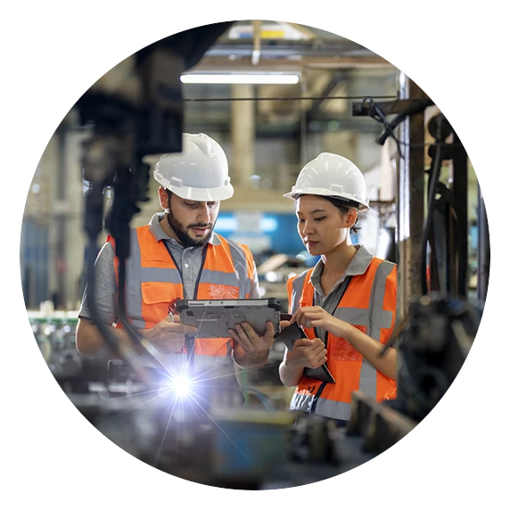 Two workers in safety vests and helmets examining a tablet in a factory.