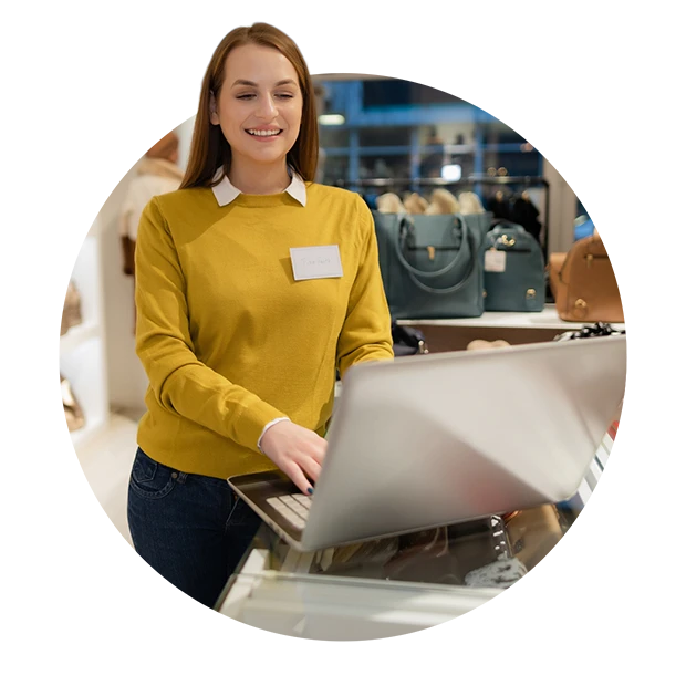 A smiling woman in a yellow sweater working on a laptop at a retail store with bags behind her.