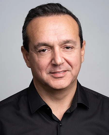A man smiling with short dark hair, wearing a black shirt against a light gray background.