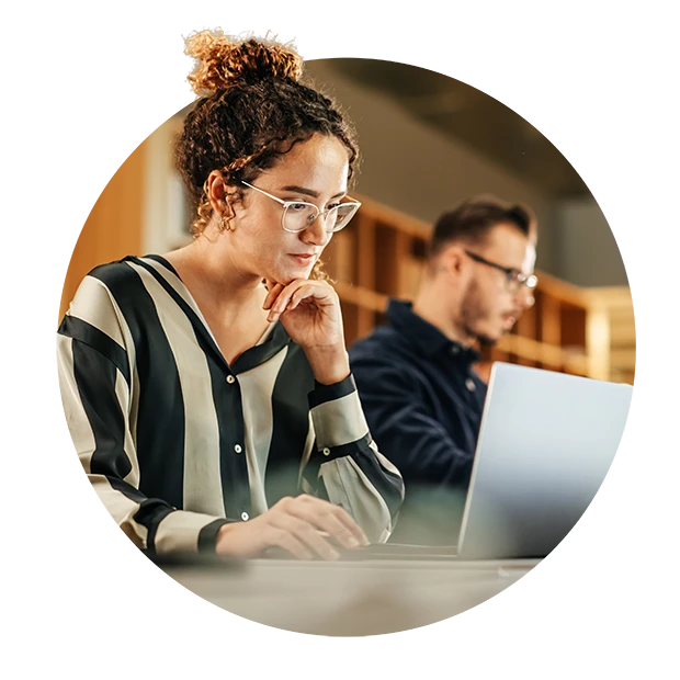 A woman wearing glasses and a striped blouse, focused on her laptop, while a man sits nearby working.