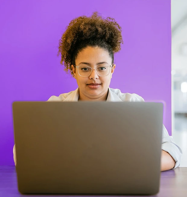 A woman with curly hair and glasses sits at a table using a laptop against a purple background.
