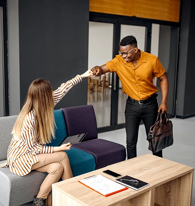 A man in an orange shirt and glasses fist bumps a woman sitting on a couch, holding a tablet.