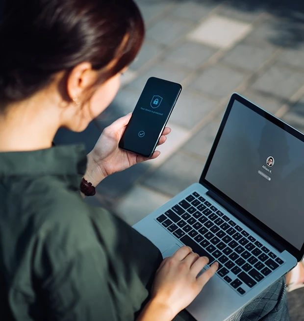 A person using a smartphone and a laptop outdoors, focusing on a login screen.