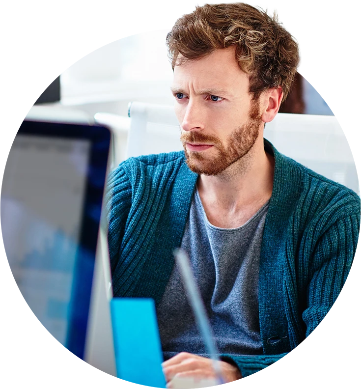 A man in a teal cardigan looks intently at a computer screen in a bright office.