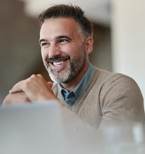 A smiling man with a beard wearing a sweater, sitting at a table, looking happy.